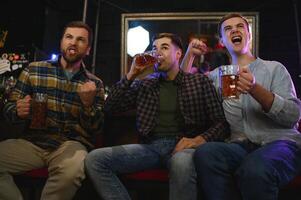 Soccer fans cheering for favourite team while enjoying drinks. Group of supporters celebrating victory while watching football match on tv in a club. Happy friends shouting while cheering for a goal. photo