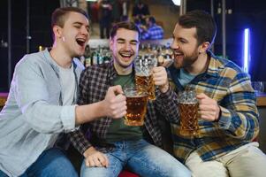 Three young men in casual clothes are smiling, holding bottles of beer while standing near bar counter in pub photo