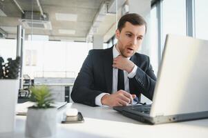 Young stressed handsome businessman working at desk in modern office shouting at laptop screen and being angry about financial situation, jealous of rival capabilities, unable to meet client needs photo