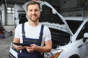Bearded mechanic in overalls standing in garage of a car salon and holding tablet. He is about to diagnostic breakdown. photo