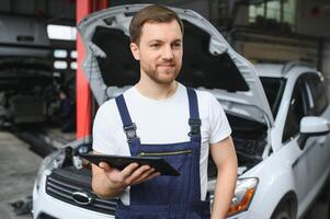 Bearded mechanic in overalls standing in garage of a car salon and holding tablet. He is about to diagnostic breakdown. photo