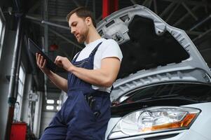 Bearded mechanic in overalls standing in garage of a car salon and holding tablet. He is about to diagnostic breakdown. photo