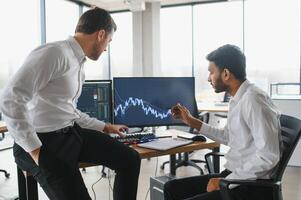 Two men traders sitting at desk at office together monitoring stocks data candle charts on screen analyzing price flow smiling cheerful having profit teamwork concept photo