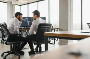 Two men traders sitting at desk at office together monitoring stocks data candle charts on screen analyzing price flow smiling cheerful having profit teamwork concept photo