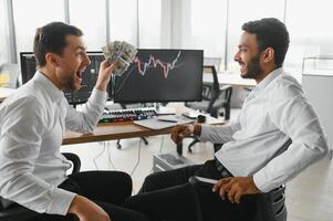 Two men traders sitting at desk at office together monitoring stocks data candle charts on screen analyzing price flow smiling cheerful having profit teamwork concept photo