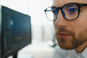 Young business man trader wearing glasses looking at computer screen with trading charts reflecting in eyeglasses watching stock trading market financial data growth concept, close up. photo