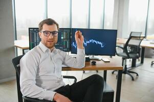 Man trader in formalwear sitting at desk in frot of monitors with charts and data at office browsing laptop checking documents analyzing stocks price changes concentrated. photo