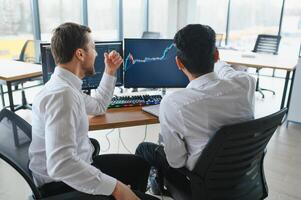 Two successful trader sitting in office and looking at screen, checking cryptocurrency information data on finance market graph, pointing on monitor photo