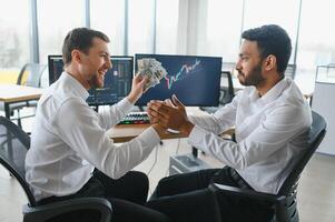 Two men traders sitting at desk at office together monitoring stocks data candle charts on screen analyzing price flow smiling cheerful having profit teamwork concept photo