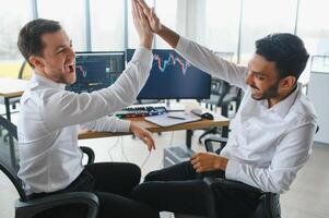 Two men traders sitting at desk at office together monitoring stocks data candle charts on screen analyzing price flow smiling cheerful having profit teamwork concept photo