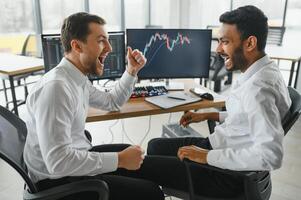 Two men traders sitting at desk at office together monitoring stocks data candle charts on screen analyzing price flow smiling cheerful having profit teamwork concept photo