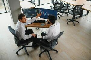 Two men traders sitting at desk at office together monitoring stocks data candle charts on screen analyzing price flow smiling cheerful having profit teamwork concept photo