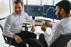 Two men traders sitting at desk at office together monitoring stocks data candle charts on screen analyzing price flow smiling cheerful having profit teamwork concept photo
