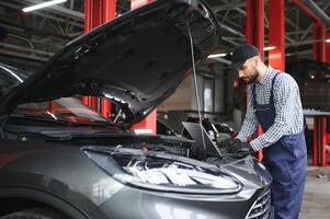 A bearded auto-mechanic in working uniform is standing next to a broken car and using the special laptop for car maintenance. An auto-mechanic in workshop repairs car. photo