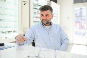 Handsome senior man choosing eyeglasses frame in optical store. photo