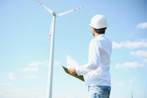 Engineer India man working at windmill farm Generating electricity clean energy. Wind turbine farm generator by alternative green energy. Asian engineer checking control electric power photo