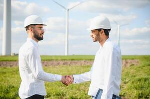 Close up of indian technician and inspector shaking hands while standing on farm with wind turbines. Concept of people, teamwork and eco friendly energy photo
