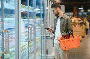 Quick text during shopping. Handsome young man holding mobile phone and smiling while standing in a food store photo