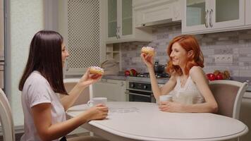 Two female friends at home relaxing and talking in the kitchen while making cheers sign with donuts. video