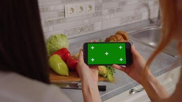 Back view of two females seaching for online recipes on a green screen chroma key horizontal phone in the kitchen. video