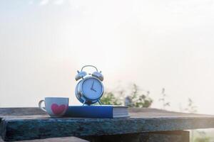 A white coffee cup and an alarm clock are placed beside the Bible on a wooden table in the morning to prepare for Bible study and watch the beautiful scenery in the morning after drinking coffee. photo