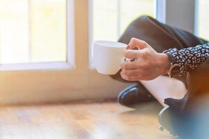 A girl sipping coffee on a wooden floor by the window in the house to see the scenery outside the house and having a coffee break before starting work at home. Continue the work from home concept photo
