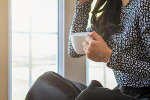 A girl sipping coffee on a wooden floor by the window in the house to see the scenery outside the house and having a coffee break before starting work at home. Continue the work from home concept photo