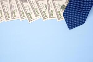 The top view of a blue tie laid on one hundred dollar banknotes isolated on a light blue background. Flat lay. Space for text. Happy Father's Day. Business and finance concept photo