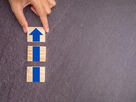Hand arranging wood block stacking with arrow up. Business growth or increasing concept picture, the rising arrow on wooden blocks laid on a dark gray stone background photo