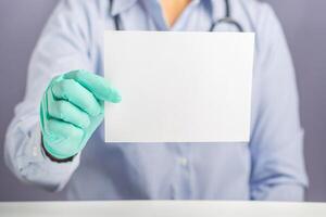 Hand of a doctor holding a blank white paper while sitting on a chair with gray background in the hospital or clinic. Close-up photo. Space for text. Medical and encouragement concept photo
