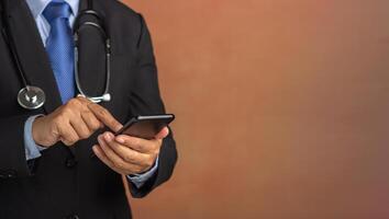 Doctor with stethoscope and dressing in a business suit using smartphone while standing with a brown background. Space for text. Medical and communication concept photo
