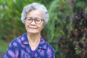 Cheerful grandmother with short gray hair wearing glasses, smiling and looking at the camera while standing in a garden. Space for text. Concept of aged people and healthcare photo