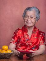 Senior woman wearing a traditional cheongsam qipao dress holding red envelopes and looking at the camera while sitting in the studio with four tangerines in a basket. Happy Chinese New Year photo