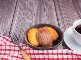 Lemon glaze madeleines in a bowl and a white coffee cup placed on a wooden table with copy space for text. Madeleine - homemade traditional French small cookie. Seashell sweet cakes photo