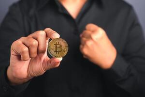 Investor guy showing a bitcoin golden coin and fist for a fight while standing in the studio with a gray background. Close-up photo. Virtual cryptocurrency and future money concept photo