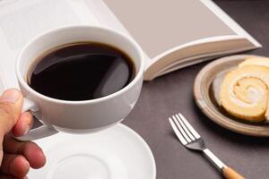 Hand holding a white coffee cup with a book, roll cakes on a plate and a cloth placed on a dark gray stone background. Close-up photo. Space for text. Concept of beverage and relaxation photo