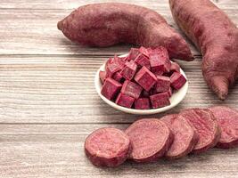 A pile of purple yams whole, sliced square in a bowl and sliced circle placed on a wooden table with copy space for text. Purple sweet potatoes. Top view. Vegan fruit and healthy fruits concept photo