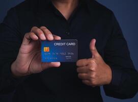 A young man holding a mockup blue credit card and showing thumb up while standing in a studio. Close-up photo. Money and business concept photo