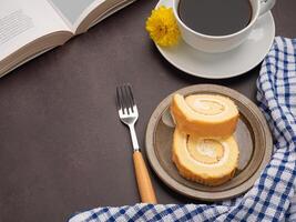 Top view of a book with roll cakes on a plate, a white coffee cup, and a cloth placed on a dark brown background. Space for text. Concept of reading and relaxation photo