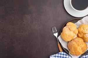 Top view of choux cream on a plate and a white coffee cup placed on a dark brown background. Space for text. Concept of relaxation photo