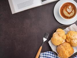 Top view of a book with choux cream on a plate and a white coffee cup placed on a dark brown background. Space for text. Concept of relaxation photo