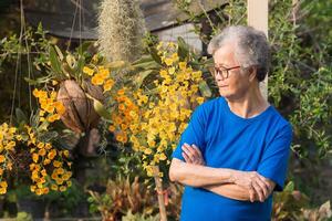 Senior woman with short gray hair wearing glasses, smiling and looking at yellow orchids while standing in a garden. Side view. Space for text. Concept of aged people and healthcare photo