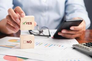 Close-up of hand holding a smartphone with the chart, graph report for the business and holding a wooden dice for choice Yes or No. Space for text. Business and planning concept photo