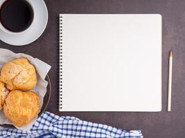 The top view of a notepad with a pencil, choux cream on a plate, and a white coffee cup are placed on dark brown background. Space for text. Concept of writing and relaxation photo