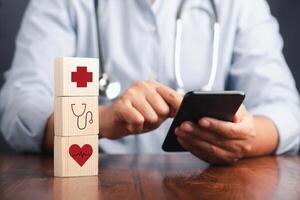 A doctor using a smartphone and wooden dice icons of health and doctors placed on the table. Close-up photo. Space for text. Concept of doctor work in maintaining human health photo
