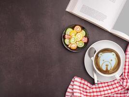 Top view of a white coffee cup with colorful sweet jelly candy on a plate and a book placed on a dark brown background. Space for text. Concept of relaxation photo