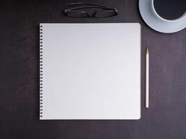 The top view of a notepad with a pencil, glasses, and a white coffee cup is placed on dark brown background. Space for text. Concept of writing and relaxation photo