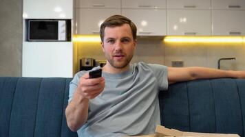 Close up of Caucasian young handsome guy watching television choosing channel while sitting on sofa and eats pizza video