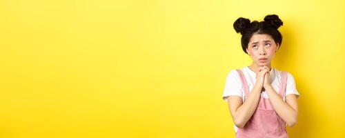 Sas asian girl holding hands in pray, pleading god, looking up and begging, standing on yellow background photo