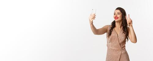 atractivo mujer en lujo vestir demostración paz firmar a teléfono cámara, tomando selfie en teléfono inteligente, posando en Navidad fiesta atuendo, en pie terminado blanco antecedentes foto